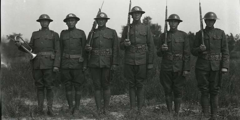 A line of 6 soldiers standing in uniform. The first soldier on the left is holding a bugle and the last four have rifles on their shoulders.