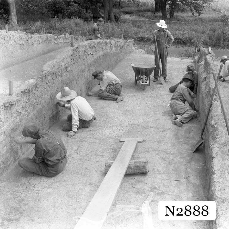 An image of the Angel Mounds Excavation. Three men on each side of the space. There is one man standing next to a wheelbarrow in the center.