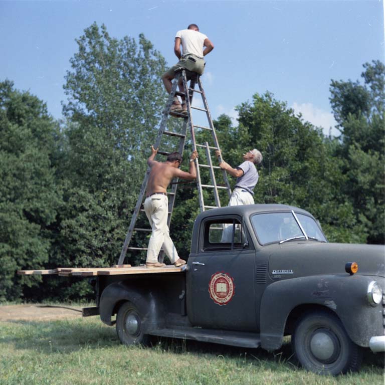 A truck with a ladder in the back. Two men are holding the ladder while a third man sits at the top of the ladder.