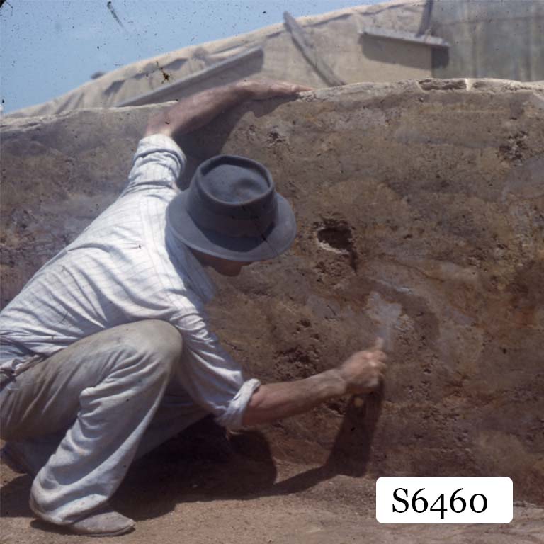 A man in a hat brushing at the wall of an excavation site.