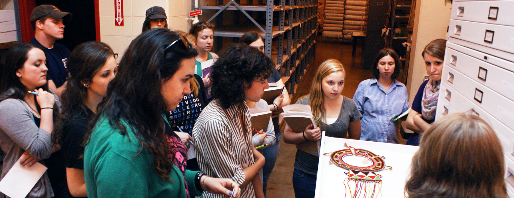 A group of students listen to an instructor speak about an artifact that is displayed on a shelf in the bottom right of the photo.